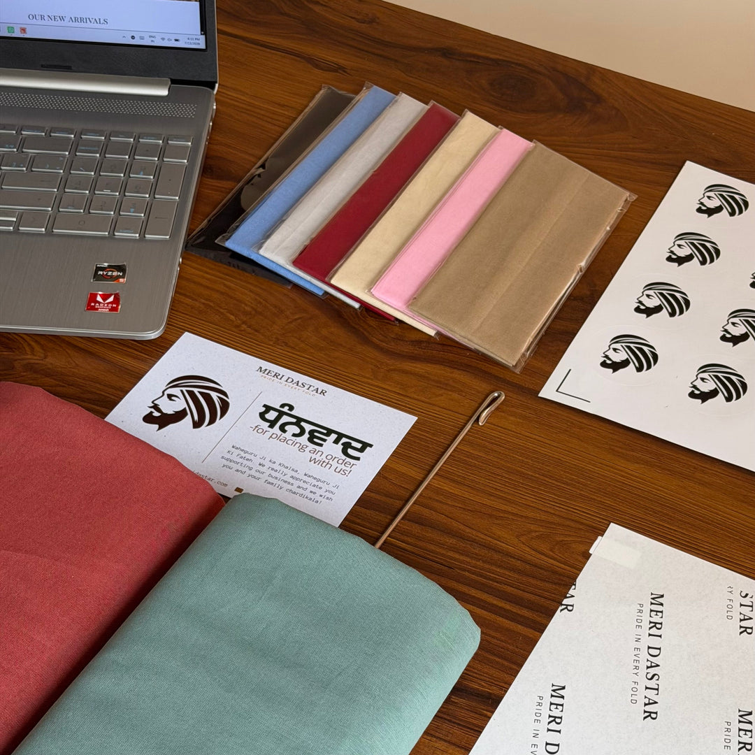 A workspace displays colorful turban fabrics, design cards, logo stickers, a laptop, and a metal turban accessory arranged on a wooden table.
