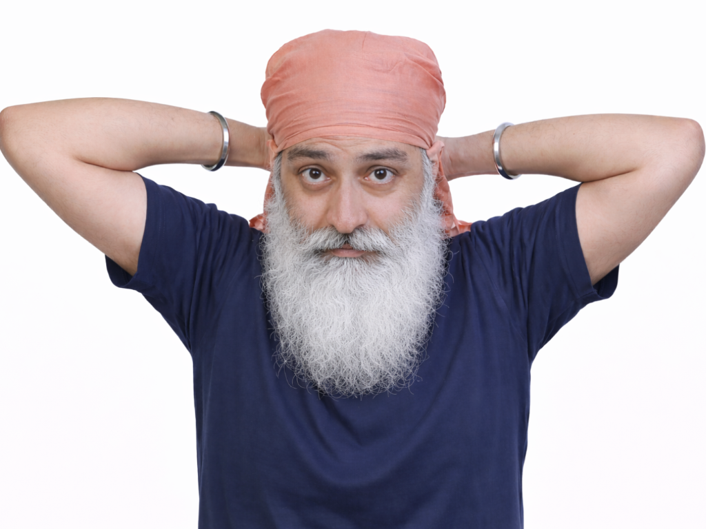 A man with a long white beard and turban base looks forward while tying fabric behind his head, arms raised to tie base of turban